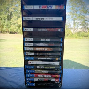 May include: A dark brown wooden game organiser filled with PS5 and PS4 video game cases. Games include "2K25", "Sniper Elite Resistance", "Alan Wake II", "Elden Ring", "Call of Duty", and "Red Dead Redemption". The organiser is on a dark surface.