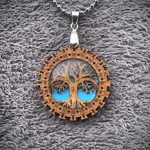 Pu&ograve; includere: Una collana con un ciondolo in legno e una catena d'argento. Il ciondolo ha un design circolare con un'immagine dell'albero della vita all'interno. L'albero &egrave; marrone con accenti blu.
