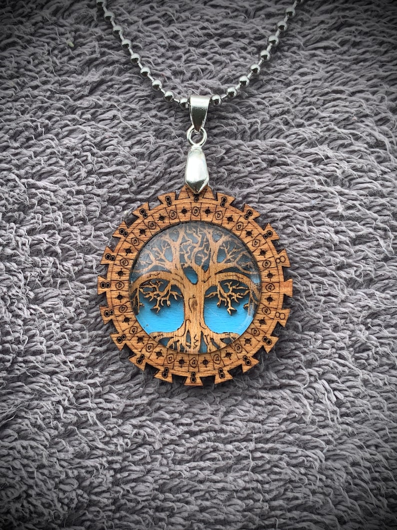 Pu&ograve; includere: Una collana con un ciondolo in legno e una catena d'argento. Il ciondolo ha un design circolare con un'immagine dell'albero della vita all'interno. L'albero &egrave; marrone con accenti blu.