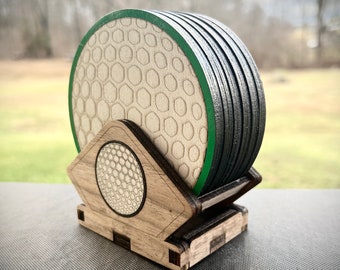 Könnte beinhalten: Satz mit sechs runden Untersetzern mit einem weißen Wabenmuster und einem grünen Rand. Die Untersetzer sind in einem Holzhalter mit einem Golfball-Design gestapelt.
