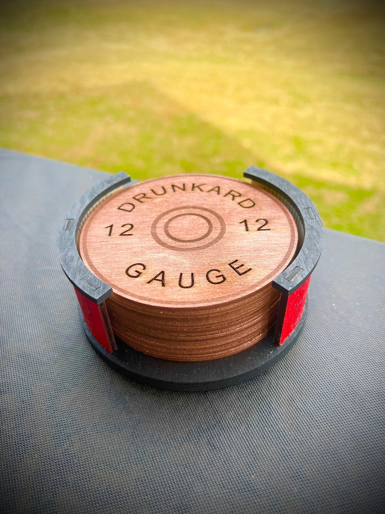 May include: A set of wooden coasters with a black and red base. The coasters are stacked and feature a brown wood grain finish. The top coaster has the text "DRUNKARD 12 GAUGE 12" engraved on it.