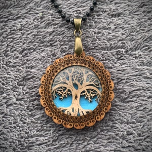 Pu&ograve; includere: Una collana con un ciondolo in legno con un design ad albero della vita. Il ciondolo &egrave; rotondo e ha uno sfondo blu con un design ad albero della vita bianco. La collana &egrave; su una catena nera.