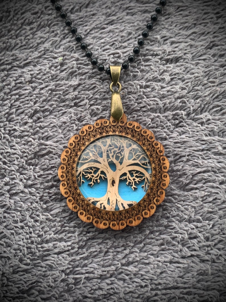 Pu&ograve; includere: Una collana con un ciondolo in legno con un design ad albero della vita. Il ciondolo &egrave; rotondo e ha uno sfondo blu con un design ad albero della vita bianco. La collana &egrave; su una catena nera.