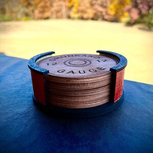 May include: A wooden coaster featuring a 12 gauge shotgun shell design. The coaster is crafted from multiple layers of wood and has a black rim. The coaster is resting on a blue surface.