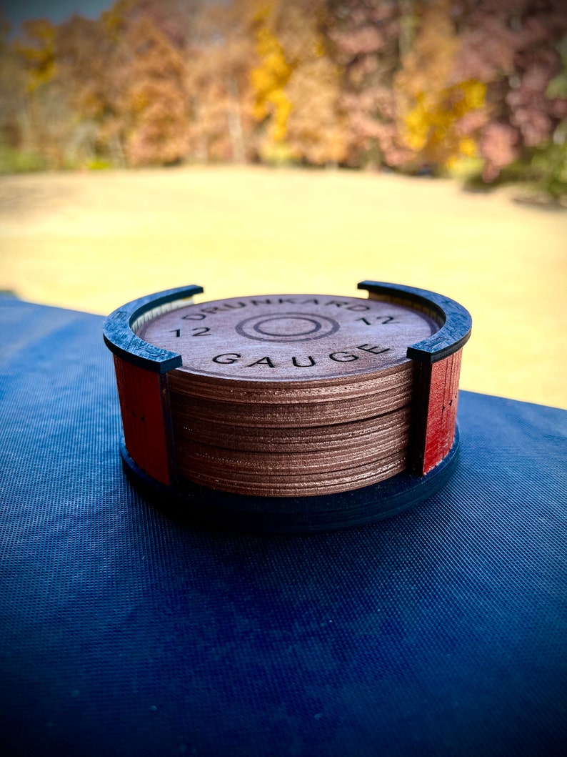 May include: A wooden coaster featuring a 12 gauge shotgun shell design. The coaster is crafted from multiple layers of wood and has a black rim. The coaster is resting on a blue surface.