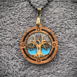 Pu&ograve; includere: Una collana con un ciondolo in legno con un design ad albero della vita. L'albero &egrave; tagliato al laser e ha uno sfondo in resina blu. Il ciondolo &egrave; attaccato a una catena di perline nere.
