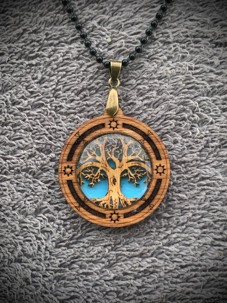 Pu&ograve; includere: Una collana con un ciondolo in legno con un design ad albero della vita. L'albero &egrave; tagliato al laser e ha uno sfondo in resina blu. Il ciondolo &egrave; attaccato a una catena di perline nere.