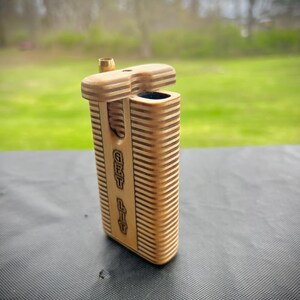 Puede incluir: Una caja de madera con una tapa con bisagras y un peque&ntilde;o tubo de metal en la parte superior. La caja est&aacute; grabada con las palabras "Get Lit".