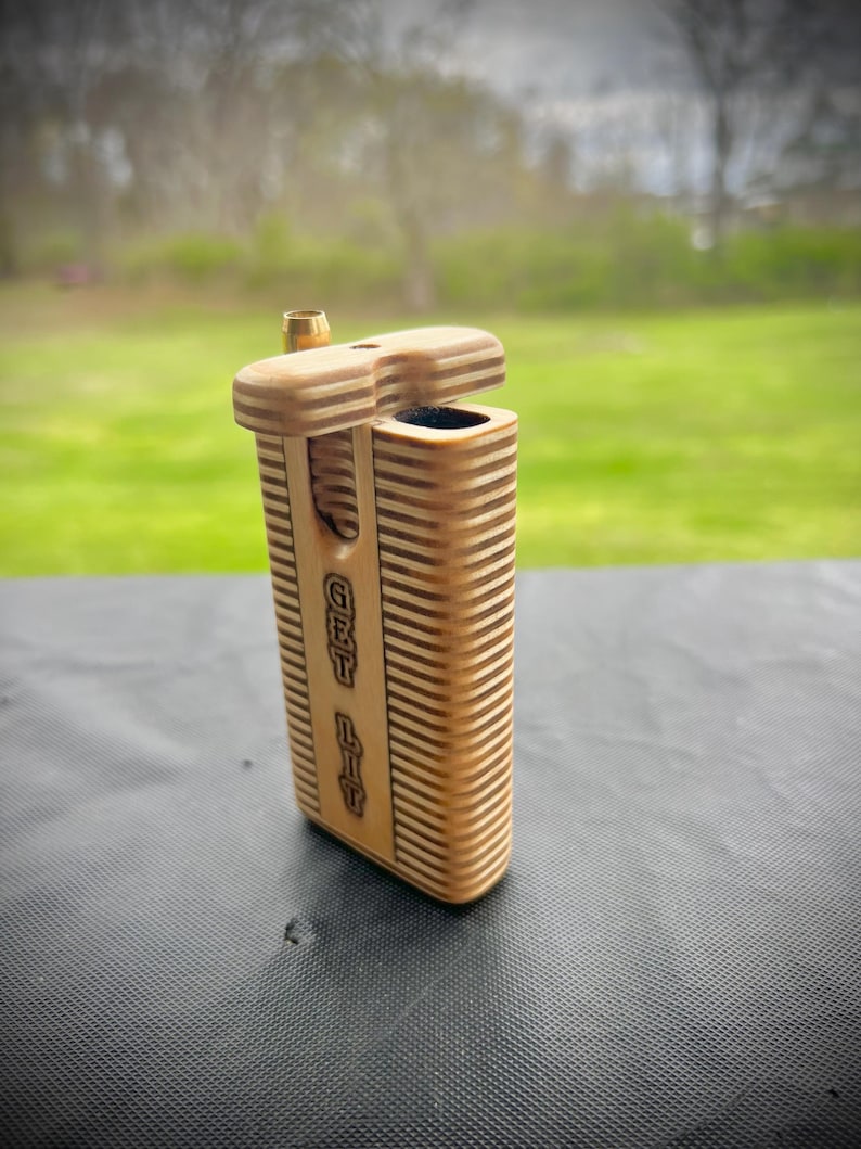 Puede incluir: Una caja de madera con una tapa con bisagras y un peque&ntilde;o tubo de metal en la parte superior. La caja est&aacute; grabada con las palabras "Get Lit".