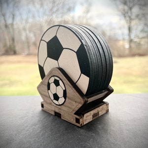 Pode incluir: Um conjunto de bases para copos com design de bola de futebol e um suporte de madeira. As bases são pretas e brancas, com um design de bola de futebol. O suporte é feito de madeira castanha clara e apresenta um gráfico de bola de futebol.