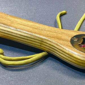 Op de afbeelding: Een houten flessenopener met een messing dop en een kleurrijk ontwerp in het midden. De opener heeft een groene paracord-koord met een messing kogel eraan.