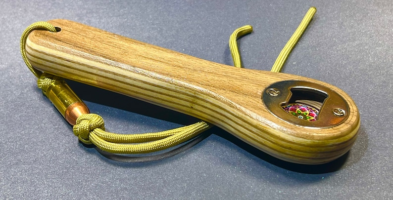 Op de afbeelding: Een houten flessenopener met een messing dop en een kleurrijk ontwerp in het midden. De opener heeft een groene paracord-koord met een messing kogel eraan.