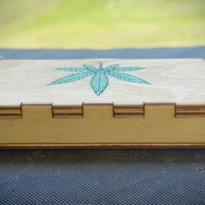 Puede incluir: Una caja de madera de color claro con tapa con bisagras. La tapa est&aacute; decorada con una hoja de cannabis turquesa. La caja tiene forma rectangular con juntas entrelazadas en los lados.