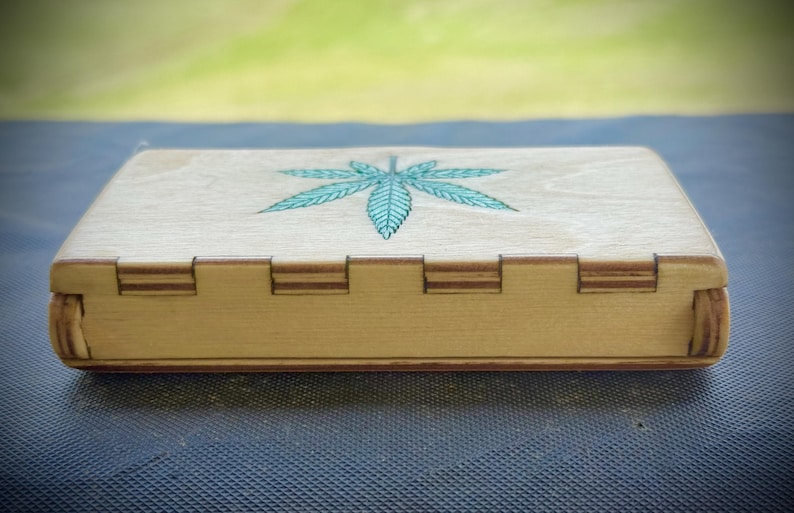 Puede incluir: Una caja de madera de color claro con tapa con bisagras. La tapa est&aacute; decorada con una hoja de cannabis turquesa. La caja tiene forma rectangular con juntas entrelazadas en los lados.