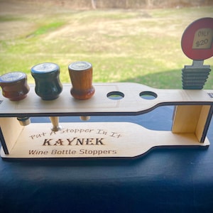Puede incluir: Soporte de madera para tapones de botellas de vino con el texto "Put a Stopper in it KAYNEK Wine Bottle Stoppers" y un cartel que dice "ONLY $20".