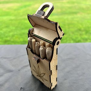 Op de afbeelding: Een houten doos met een scharnierend deksel, lasergesneden met een bladeren ontwerp. De doos is open en onthult verschillende gerolde sigaretten. De doos heeft een slotvormige handgreep. De achtergrond is een vage groene buitenscène.