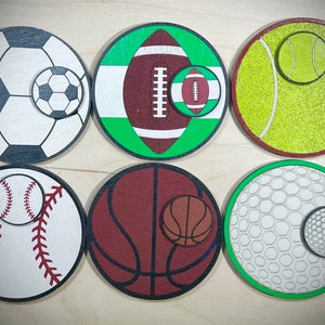 May include: Six round wooden coasters with different sports ball designs. Each coaster features a larger ball design with a smaller version of the same ball in the corner. The coasters feature a soccer ball, football, tennis ball, baseball, basketball, and golf ball.
