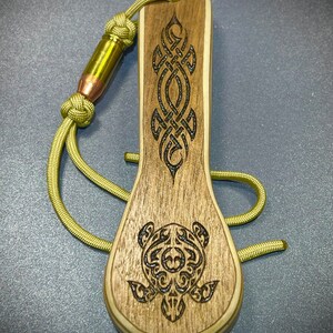 Op de afbeelding: Een houten flessenopener met een bruin en zwart schildpadontwerp en een Keltisch knoopontwerp. De opener is bevestigd aan een groene paracord-koord met een kogelhulsel.
