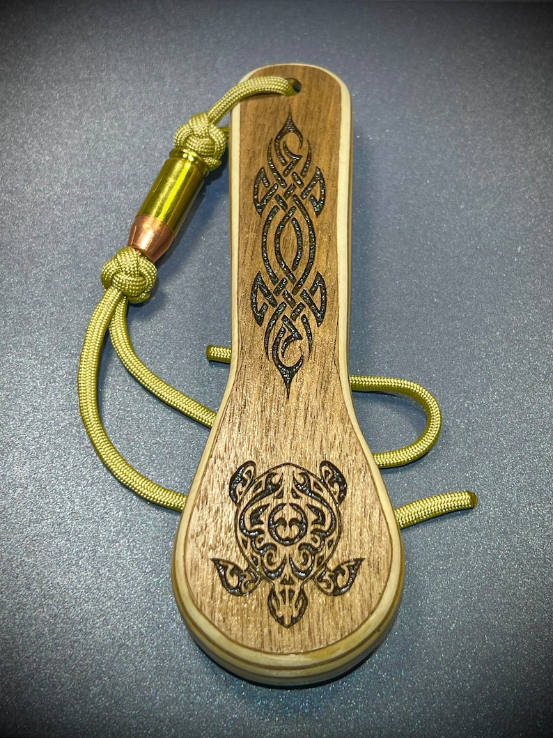 Op de afbeelding: Een houten flessenopener met een bruin en zwart schildpadontwerp en een Keltisch knoopontwerp. De opener is bevestigd aan een groene paracord-koord met een kogelhulsel.