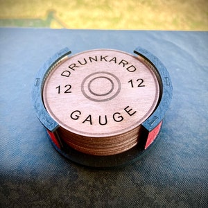 May include: A set of six brown wooden coasters with a black and red holder. The coasters are round and have the words "Drunkard 12 Gauge" engraved on them.