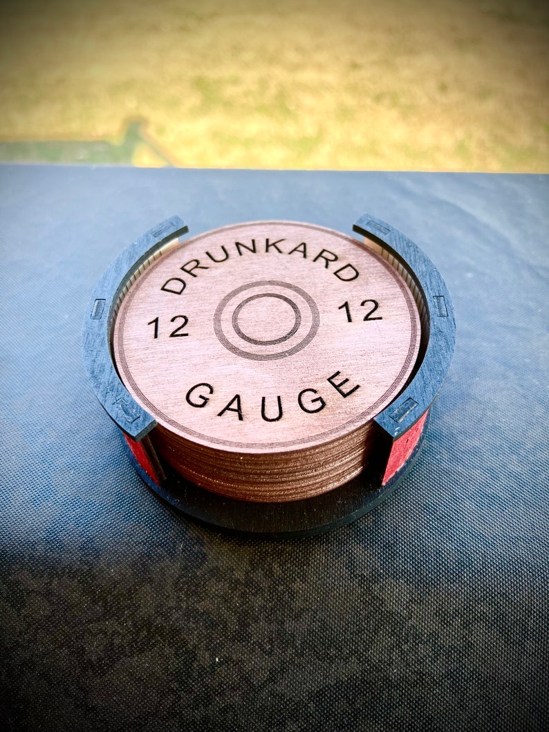 May include: A set of six brown wooden coasters with a black and red holder. The coasters are round and have the words "Drunkard 12 Gauge" engraved on them.