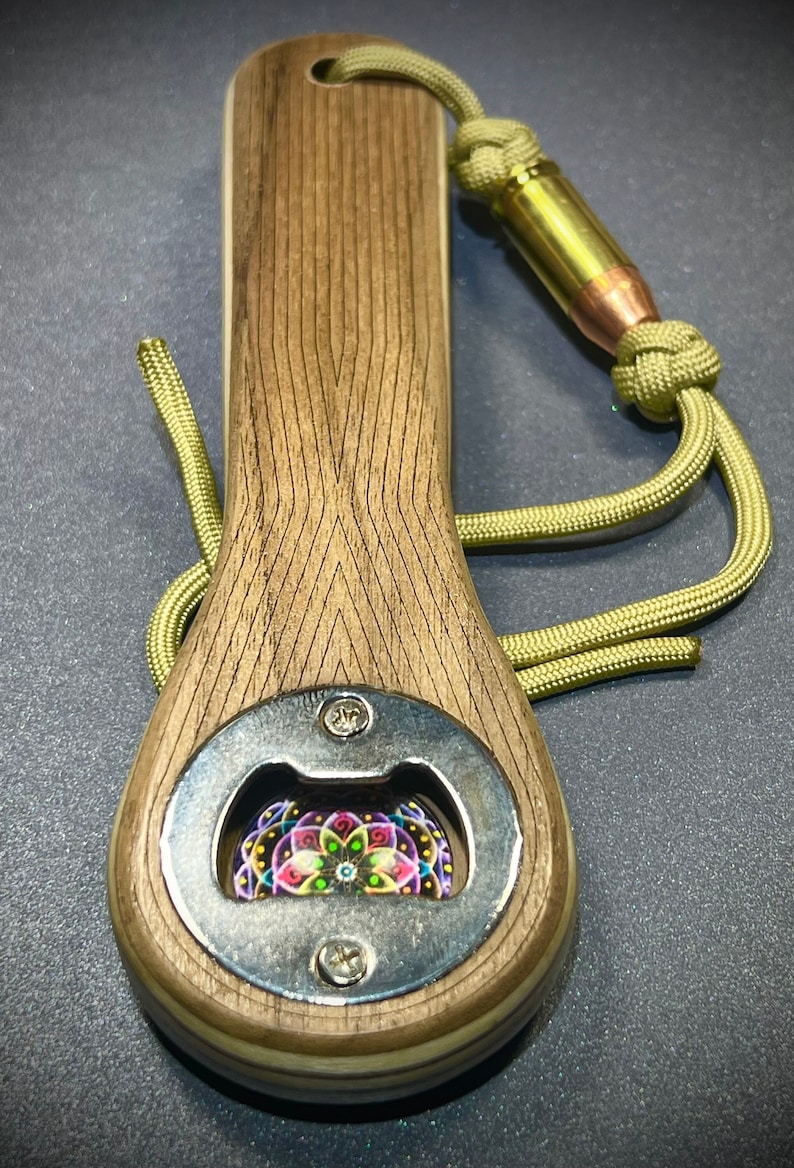 Op de afbeelding: Een houten flessenopener met een metalen dopopener en een kleurrijk mandala-ontwerp in het midden. De opener heeft een groene paracord-koord met een messing kogelaccent.