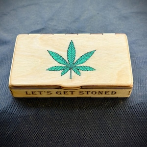 May include: A rectangular wooden box with a hinged lid. The lid features a green cannabis leaf design. The phrase "LET'S GET STONED" is printed on the front of the box. The box is made of light-coloured wood.