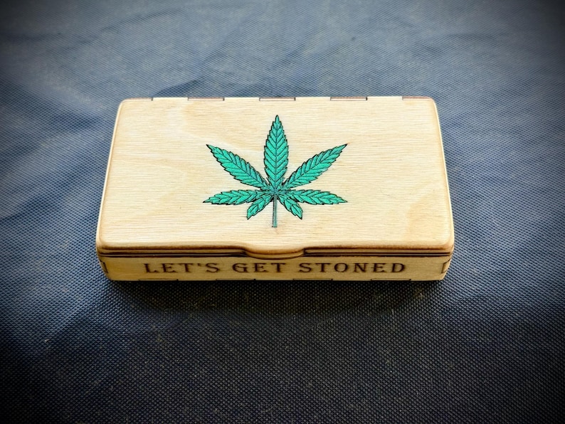 Puede incluir: Caja rectangular de madera con tapa con bisagras. La tapa presenta un dise&ntilde;o de hoja de cannabis verde. La frase "LET'S GET STONED" est&aacute; impresa en la parte delantera de la caja. La caja es de madera clara.