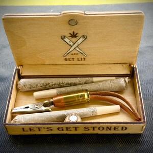 Puede incluir: Caja de madera con el texto "LET'S GET STONED" y "GET LIT" con una hoja de cannabis y porros cruzados. Contiene cigarrillos liados, una pinza met&aacute;lica y un accesorio con forma de bala con una correa de cuero.