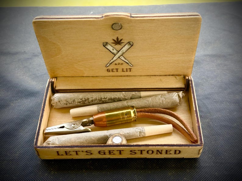 Puede incluir: Caja de madera con el texto "LET'S GET STONED" y "GET LIT" con una hoja de cannabis y porros cruzados. Contiene cigarrillos liados, una pinza met&aacute;lica y un accesorio con forma de bala con una correa de cuero.