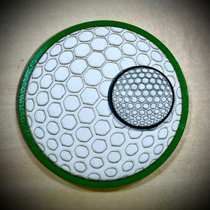 Puede incluir: Un posavasos redondo de madera verde con un dise&ntilde;o de bola de golf blanco y verde y un dise&ntilde;o de bola de golf m&aacute;s peque&ntilde;o en el centro.