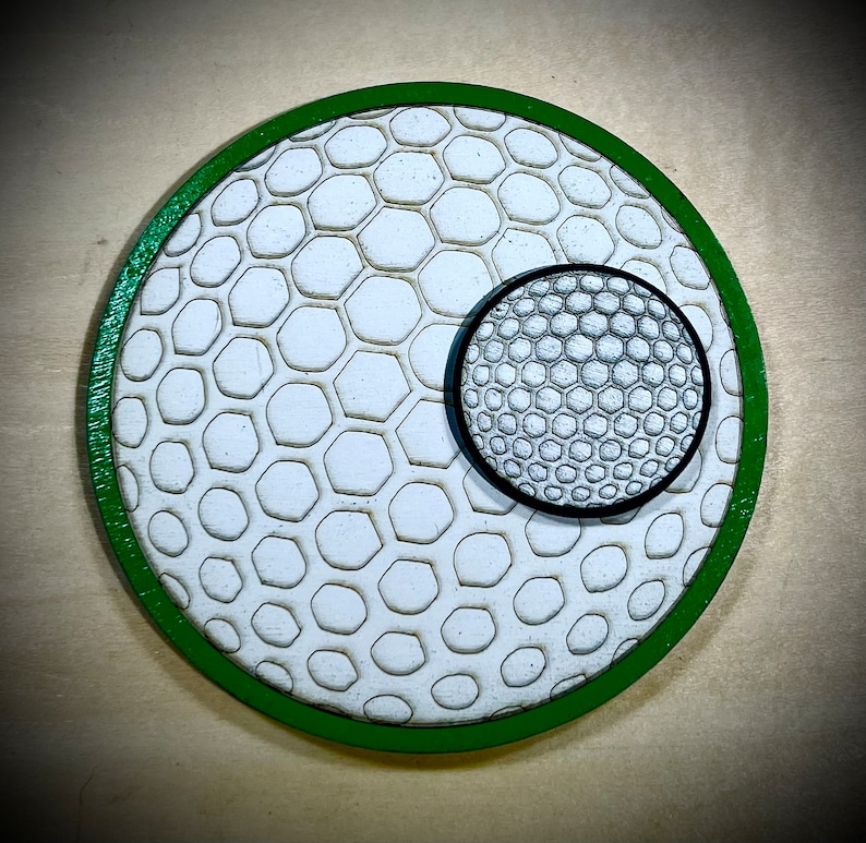 Puede incluir: Un posavasos redondo de madera verde con un dise&ntilde;o de bola de golf blanco y verde y un dise&ntilde;o de bola de golf m&aacute;s peque&ntilde;o en el centro.