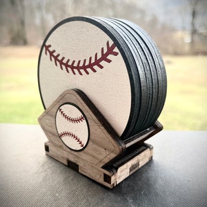 May include: A set of six wooden coasters featuring a baseball design. The coasters are stacked in a holder shaped like a baseball diamond. The coasters are made of wood and have a white background with a red baseball stitch design. The holder is made of wood and has a brown stain.