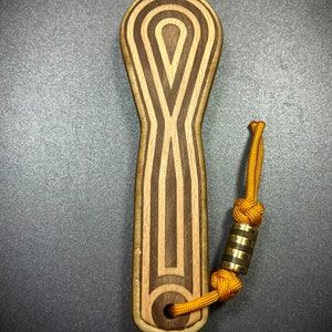 Op de afbeelding: Een houten flessenopener met een bruin en licht houtpatroon. De opener heeft een lus aan de bovenkant om aan een sleutelhanger te bevestigen. De lus is gemaakt van een geel koord met een gouden metalen kraal.