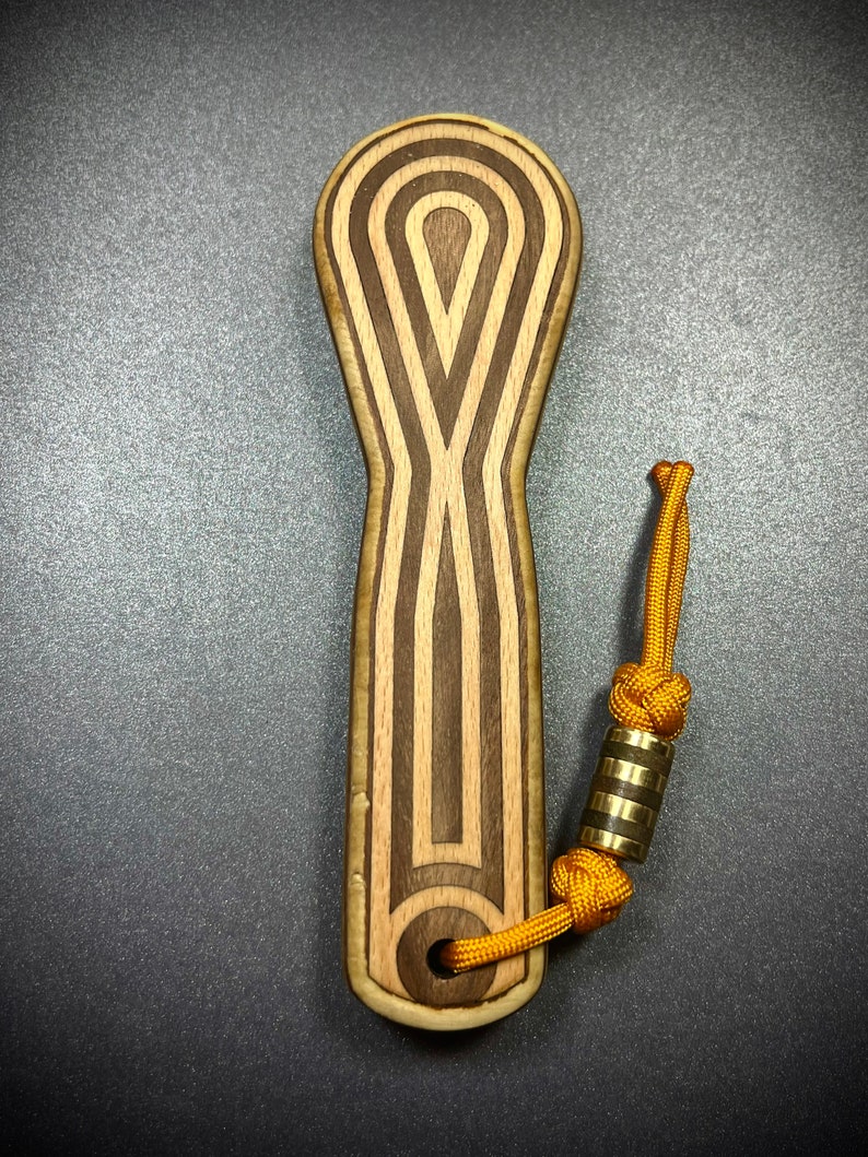 Op de afbeelding: Een houten flessenopener met een bruin en licht houtpatroon. De opener heeft een lus aan de bovenkant om aan een sleutelhanger te bevestigen. De lus is gemaakt van een geel koord met een gouden metalen kraal.