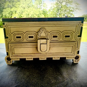 May include: A wooden decorative box with intricate geometric patterns and a latch closure. The box is light-colored with dark accents and sits on small, rounded feet. The background is a blurred outdoor scene.