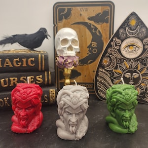 May include: Three hand-crafted candles shaped like Medusa's head. The candles are red, gray, and green. The candles are sitting on a black surface in front of a wooden tarot card, a skull, and a wooden Ouija board.
