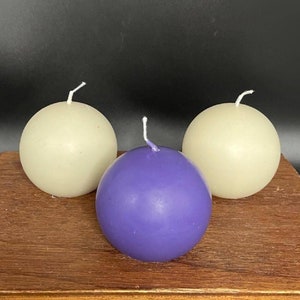 May include: Three round candles, two are off-white and one is purple. The candles are on a wooden surface.