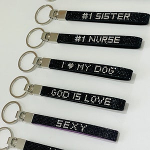Pode incluir: Sete chaveiros pretos com argolas prateadas. Cada chaveiro tem uma frase diferente em strass: #1 Mom, #1 Sister, #1 Nurse, I Love My Dogs, God Is Love, Sexy, XOXO.