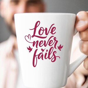 Puede incluir: Taza de cerámica blanca con la frase "Love never fails" en borgoña, en escritura cursiva. El diseño incluye un corazón e ilustraciones de pájaros. La taza está sujeta por una persona.