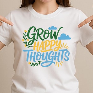 Puede incluir: Camiseta blanca con la frase "Grow Happy Thoughts" en verde, amarillo y azul. El diseño incluye elementos florales y nubes. La camiseta es de cuello redondo.