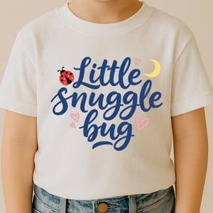 Puede incluir: Camiseta blanca con la frase "Little snuggle bug" en escritura azul, una mariquita roja, corazones rosas y una luna creciente amarilla. Prenda infantil.