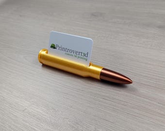 Può includere: Un porta biglietti da visita a forma di proiettile color oro con punta color rame. Un biglietto da visita bianco è inserito nel supporto, con il testo "Printrovert3d custom 3d printing". Il supporto è su una superficie di legno.