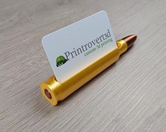 Può includere: Un porta biglietti da visita dorato a forma di bossolo di proiettile con un biglietto da visita bianco che recita "Printrovert3d stampa 3d personalizzata".