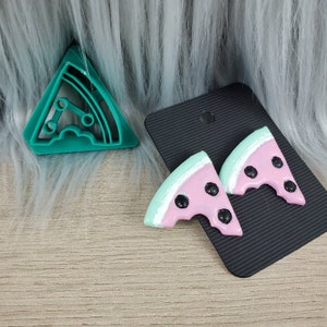 May include: A pair of watermelon slice earrings with black seeds. The earrings are pink and green and have a bite taken out of them. The earrings are on a black background.