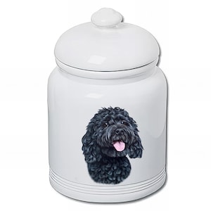 Peut inclure: Un pot en céramique blanc avec un couvercle, orné d'un portrait de chien noir. Le chien a des poils bouclés et une langue rose. Le pot est de forme ronde avec des rayures noires près de la base.