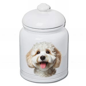 Peut inclure: Pot à friandises pour animaux de compagnie en céramique blanche avec couvercle. Le pot présente une image réaliste et souriante d'un chien duveteux de couleur crème avec une langue rose. Le pot a des lignes horizontales noires près de la base.