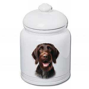 May include: White ceramic jar with a lid, decorated with a detailed portrait of a brown and black dog. The dog has a long snout, floppy ears, and a happy expression. The jar has black horizontal stripes near the base.