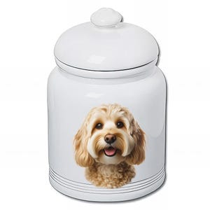 Peut inclure: Pot à friandises pour animaux de compagnie en céramique blanche avec couvercle. Le pot présente une image détaillée en couleur d'un chien Cavapoo brun clair souriant. Le pot a des lignes horizontales noires près de la base.