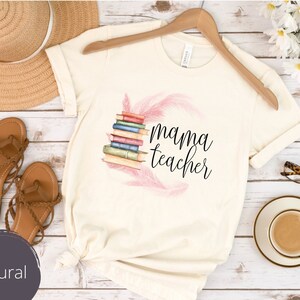 May include: A white t-shirt with a pink feather design and the text "mama teacher" in black. The shirt is laying on a wooden surface with a straw hat, sandals, and a cup of coffee.